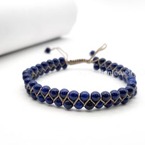May include: A double-strand bracelet featuring deep blue, spherical beads. The beads are strung on a beige cord, creating a woven pattern. The bracelet has adjustable cords for a custom fit. The jewelry is displayed on a white surface.
