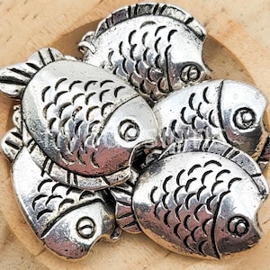 May include: A collection of silver-colored fish-shaped charms. Each charm has detailed scales and fins, with a small eye visible on each fish. The charms are arranged in a pile, creating a textured and metallic appearance. These charms could be used for jewelry making or other craft projects.