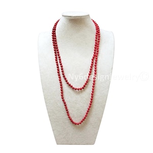 May include: A long, red beaded necklace draped on a white mannequin. The necklace is composed of small, round beads and is displayed in a layered style. The beads are a vibrant, solid red color.