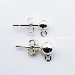 May include: Two silver-colored stud earrings with a ball design. Each earring has a post for insertion and a small loop for attaching charms or other decorative elements. The earrings are displayed against a white background.