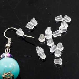 May include: A close-up of a set of clear plastic earring backs. The backs are small and round, with a slight indentation in the centre. There are 15 backs in total, and they are scattered on a black surface.