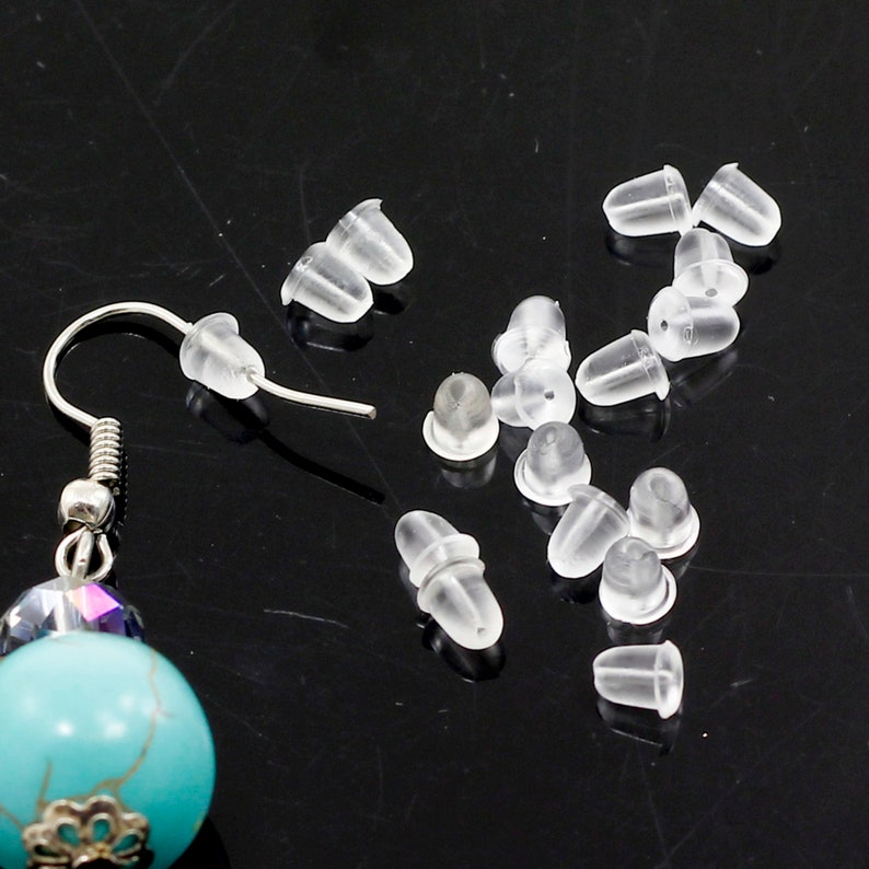 May include: A close-up of a set of clear plastic earring backs. The backs are small and round, with a slight indentation in the centre. There are 15 backs in total, and they are scattered on a black surface.