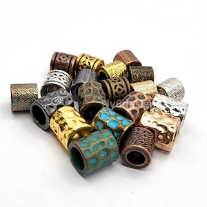 May include: A collection of cylindrical metal beads in various finishes including antique copper, polished gold, silver, and dark bronze. The beads feature diverse surface textures such as hammered, cross-hatched, and intricate cut-out patterns.