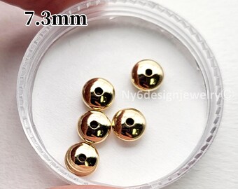 May include: A collection of five small, gold-colored beads, each with a central hole, displayed in a clear plastic container. The beads are approximately 7.3mm in diameter. The image is well-lit, highlighting the metallic sheen of the beads.