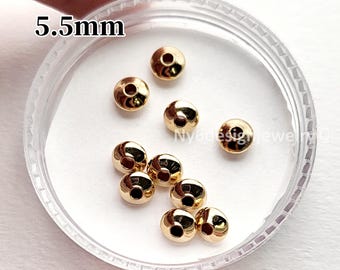 May include: A collection of small, gold-colored beads, each with a 5.5mm diameter, are displayed inside a clear, round container. The beads have a rounded shape with a hole through the center. The text "5.5mm" is visible in the upper left corner.