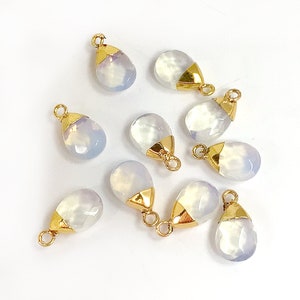 May include: A collection of teardrop-shaped pendants. Each pendant features a faceted, pale blue-white gemstone with a gold-colored metal cap and loop. The pendants are scattered on a white surface, showcasing their delicate design.