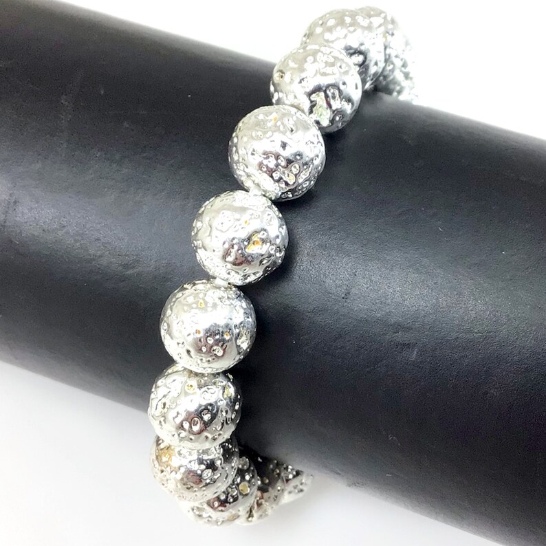 May include: A silver bracelet featuring large, textured, spherical beads. The beads have a hammered metal appearance, reflecting light. The bracelet is displayed against a black background, highlighting its metallic sheen. The jewelry is a fashion accessory.