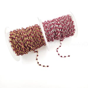 May include: Two spools of beaded chain. One spool features a gold-colored chain with small, dark red beads. The other spool has a silver-colored chain with similar dark red beads. The spools are on a white background.