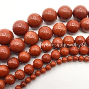 May include: Close-up of strands of red jasper gemstone beads in various sizes. The beads are round and have a polished, glossy finish. The color is a deep, earthy red with natural variations. The image includes the text "Ny6designjewelry©".