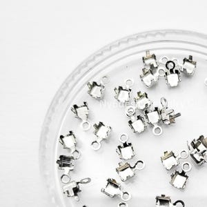 May include: A collection of small, silver-colored jewelry findings, scattered inside a clear, round container. Each finding has a square top with four prongs and a small loop at the bottom. The findings are designed for jewelry making.