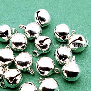 May include: A close-up of a group of silver jingle bells. The bells are round and have a small loop at the top for attaching. They are scattered on a green surface.