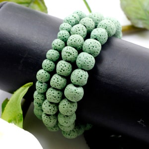 May include: A multi-strand bracelet composed of round, light green porous beads with a textured, pitted surface. The beads are strung together in a circular arrangement, showcasing a consistent matte finish and a uniform pale green color throughout.