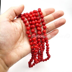 May include: A strand of bright red, round beads. The beads are held in a hand against a white background. The beads are strung together, forming a necklace or bracelet.