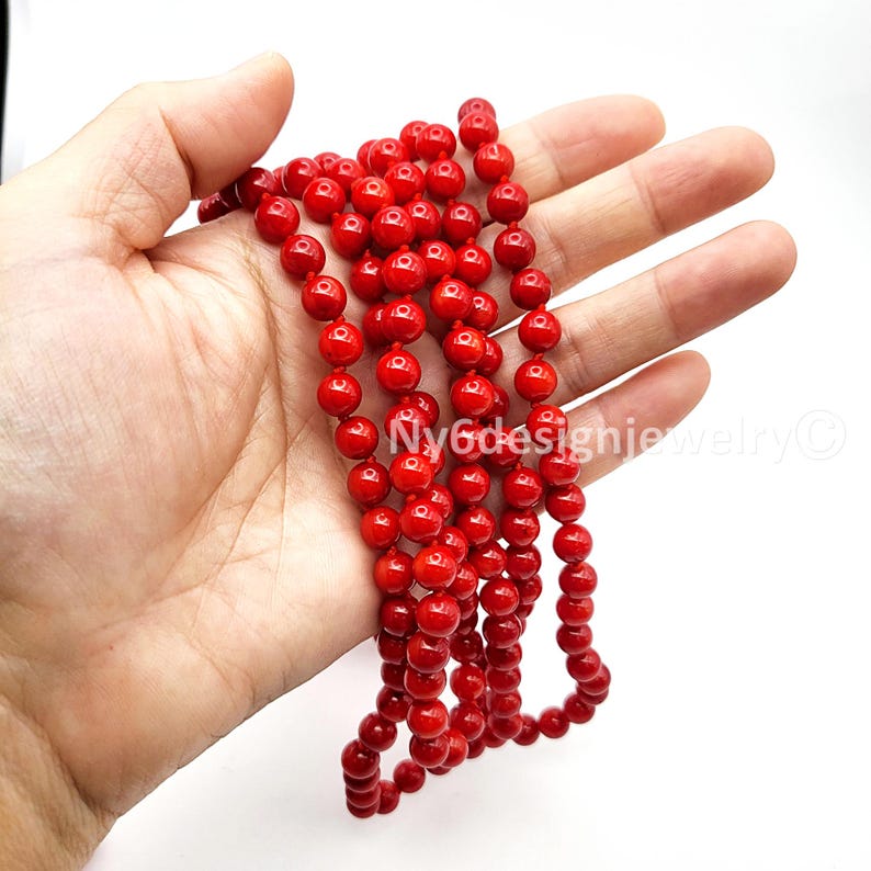 May include: A strand of bright red, round beads. The beads are held in a hand against a white background. The beads are strung together, forming a necklace or bracelet.
