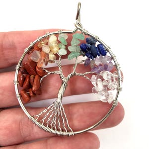 May include: A silver wire wrapped tree of life pendant with seven different colored gemstones. The stones are arranged in a circle around the tree, with red at the bottom and purple at the top. The pendant is approximately 2 inches in diameter.