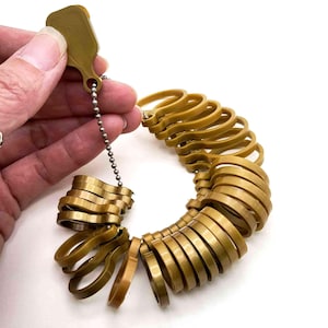 May include: A set of gold-colored plastic ring sizers connected by a metal ball chain. The collection includes multiple circular bands of varying diameters, each featuring a flat tab with a hole for the chain, designed for measuring finger size.