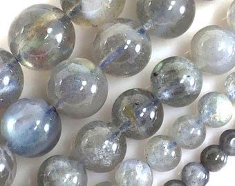 May include: Close-up of a strand of round, iridescent labradorite beads. The beads are primarily gray with flashes of blue, green, and gold. The beads are strung together, showcasing their polished, smooth surface. Ideal for jewelry making.