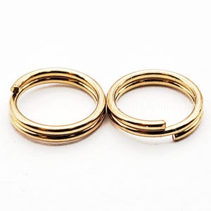May include: Two gold-colored split key rings. Each ring is made of three parallel bands. The rings are open, ready to receive keys or other items. The background is plain white.