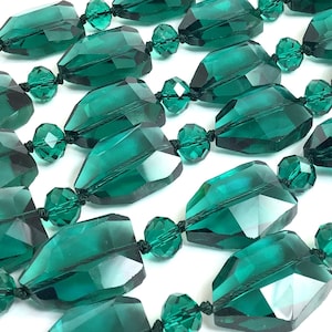 May include: Close-up of a necklace featuring large, faceted, emerald-green beads. The beads are strung together with black string, creating a repeating pattern. Smaller, round beads separate the larger, geometric beads. The background is white.