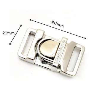May include: A silver-colored metal buckle with the text "Me & Mine". The buckle is 40mm long and 21mm wide. It has a hinged design with a central pivoting piece and two rectangular loops.