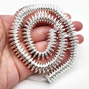 May include: A long strand of shiny silver-colored bicone-shaped beads arranged in a spiral pattern. Each bead features a pointed center and a smooth, reflective metallic finish. The beads are strung together on a thin, light-colored cord.