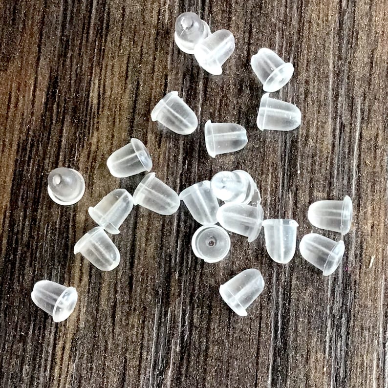 May include: A close-up of a group of clear plastic earring backs. The backs are small and round, with a slight indentation in the centre. They are scattered on a wooden surface.