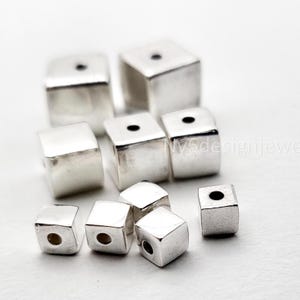 May include: A collection of silver-colored, cube-shaped beads. Each bead has a hole drilled through the center, and the surface is smooth and reflective. The beads are scattered on a white surface, with some in focus and others blurred.