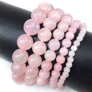 May include: Set of five pink rose quartz bracelets with varying bead sizes. The bracelets are stacked on a black surface.