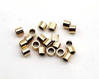May include: A collection of small, cylindrical gold-colored metal beads scattered on a white surface. Each bead has a hole running through the center, and they appear to be uniform in size and shape. These beads are likely used for jewelry making or other craft projects.