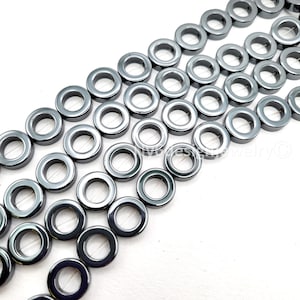 May include: Rows of dark gray, donut-shaped beads with a metallic sheen. The beads have a circular hole in the center and are strung together, creating a pattern. These beads are suitable for jewelry making and crafting projects.