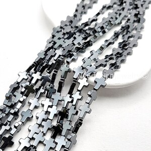 May include: Strands of gray cross-shaped beads. The beads are strung together, creating a chain-like appearance. The crosses have a metallic sheen and are arranged in a diagonal pattern against a white background. The beads are likely for jewelry making.