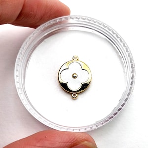 May include: A gold-toned charm with a white clover design, set within a clear plastic container. The charm features a four-leaf clover shape with a small central dot. The charm has two small loops for attachment.