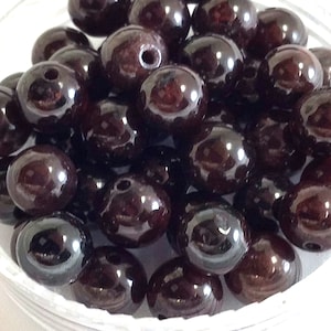 May include: A close-up of a bowl filled with dark red, oval-shaped beads. The beads are smooth and shiny, with a slight metallic sheen.