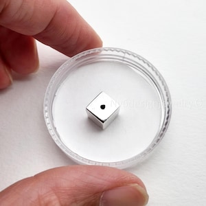 May include: A small, silver-colored die with a single black dot, resting inside a clear, round container. The die is square-shaped and appears to be made of metal. The container sits on a white surface.