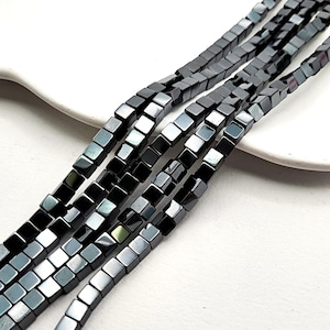 May include: Strands of square, dark gray hematite beads. The beads are strung together, forming multiple lines of the same material. The beads have a metallic sheen and are arranged on a white surface.