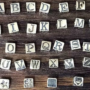 May include: A set of 26 silver letter beads, each with a different letter of the alphabet, plus three additional beads with a star, a flower, and a heart design. The beads are arranged in rows on a wooden surface.