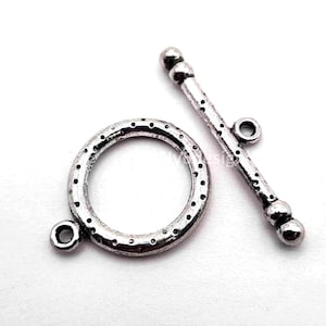 May include: A silver-toned metal toggle clasp for jewelry making, consisting of a circular ring with a dotted texture and a matching T-bar component. The ring features a small loop for attachment, while the T-bar has rounded ends and a central loop.