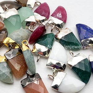 May include: A collection of gemstone pendants in various colors, including green, red, and blue. Each pendant has a silver or gold-colored metal cap and a loop for attaching to a chain or cord. The pendants are faceted and have a polished finish.