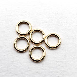 May include: Five small, gold-colored, circular jewelry findings are scattered on a white surface. Each ring has a smooth, polished finish and a consistent thickness. These findings are suitable for jewelry making.