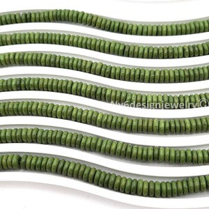 May include: Close-up of several strands of olive green wooden beads. The beads are small, round, and strung together, creating a wavy pattern across a white surface. The beads are evenly spaced and have a natural, earthy appearance.