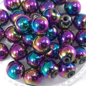 May include: A collection of iridescent round beads in shades of purple, blue, and green. The beads have a metallic finish and are scattered on a white surface.