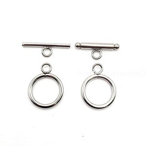 May include: Two silver-colored toggle clasps for jewelry making. Each clasp consists of a ring and a bar. One bar is a straight rod, and the other has a ball on each end. The clasps are on a white background.