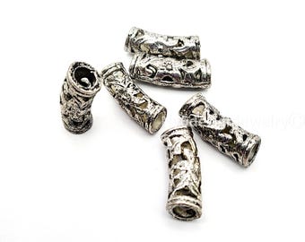 May include: Six silver-colored, cylindrical beads with intricate, openwork designs. The beads have a distressed, antique finish and are scattered on a white background. These decorative beads are suitable for jewelry making.