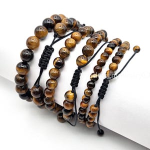 May include: Several adjustable bracelets featuring tiger eye beads in varying sizes. The beads display a mix of brown and gold tones. Each bracelet has a black, woven cord with a sliding knot closure for a customizable fit.