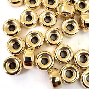 Pu&ograve; includere: Un gruppo di perline in metallo dorate con un anello rialzato attorno al foro centrale. Le perline sono sparse su una superficie bianca.