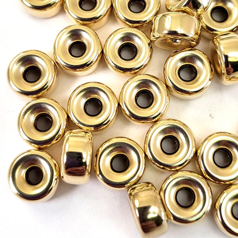 Pu&ograve; includere: Un gruppo di perline in metallo dorate con un anello rialzato attorno al foro centrale. Le perline sono sparse su una superficie bianca.
