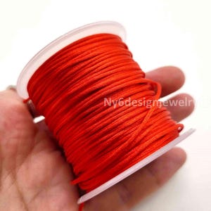 May include: A spool of vibrant red cord wrapped tightly around a translucent plastic cylinder. The cord has a smooth, consistent texture and is wound in multiple layers. The text Ny6designjewelry is visible on the surface of the red cord.
