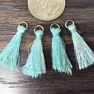 May include: Four turquoise tassels with silver accents and small metal rings. The tassels are arranged near a quarter coin, showcasing their size and detail. The tassels are made of a soft, silky material.