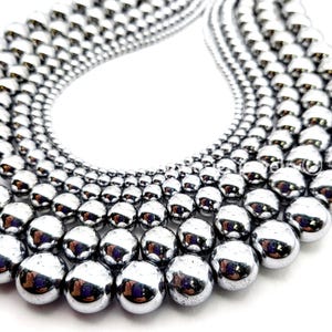 May include: Close-up of multiple strands of shiny, silver-colored beads. The beads are spherical and reflect light, creating a bright, metallic appearance. The beads vary in size, with larger beads in the foreground and smaller ones receding into the background. The beads are likely for jewelry making.