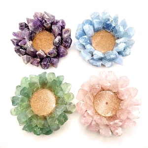 May include: Four circular candle holders arranged in a square formation. Each holder features a central circular cork base surrounded by a ring of raw, jagged crystal clusters in purple, light blue, pale green, and soft pink colors respectively.